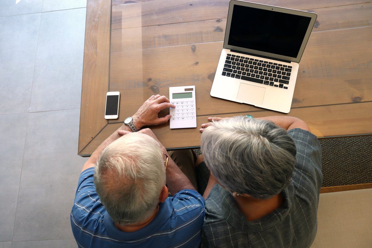 Homem e mulher idosos, de cabelos brancos, sentados em uma mesa em frente a um computador, calculadora e celular, vistos de cima