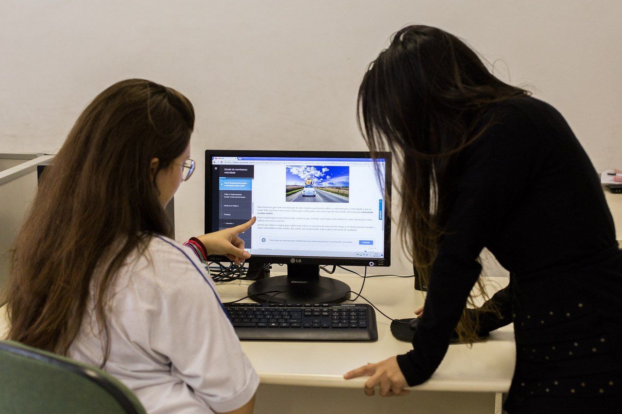 Duas mulheres de costas, uma de pé e outra sentada, olham a tela de um computador (Crédito: Diogo Moreira/A2 FOTOGRAFIA)