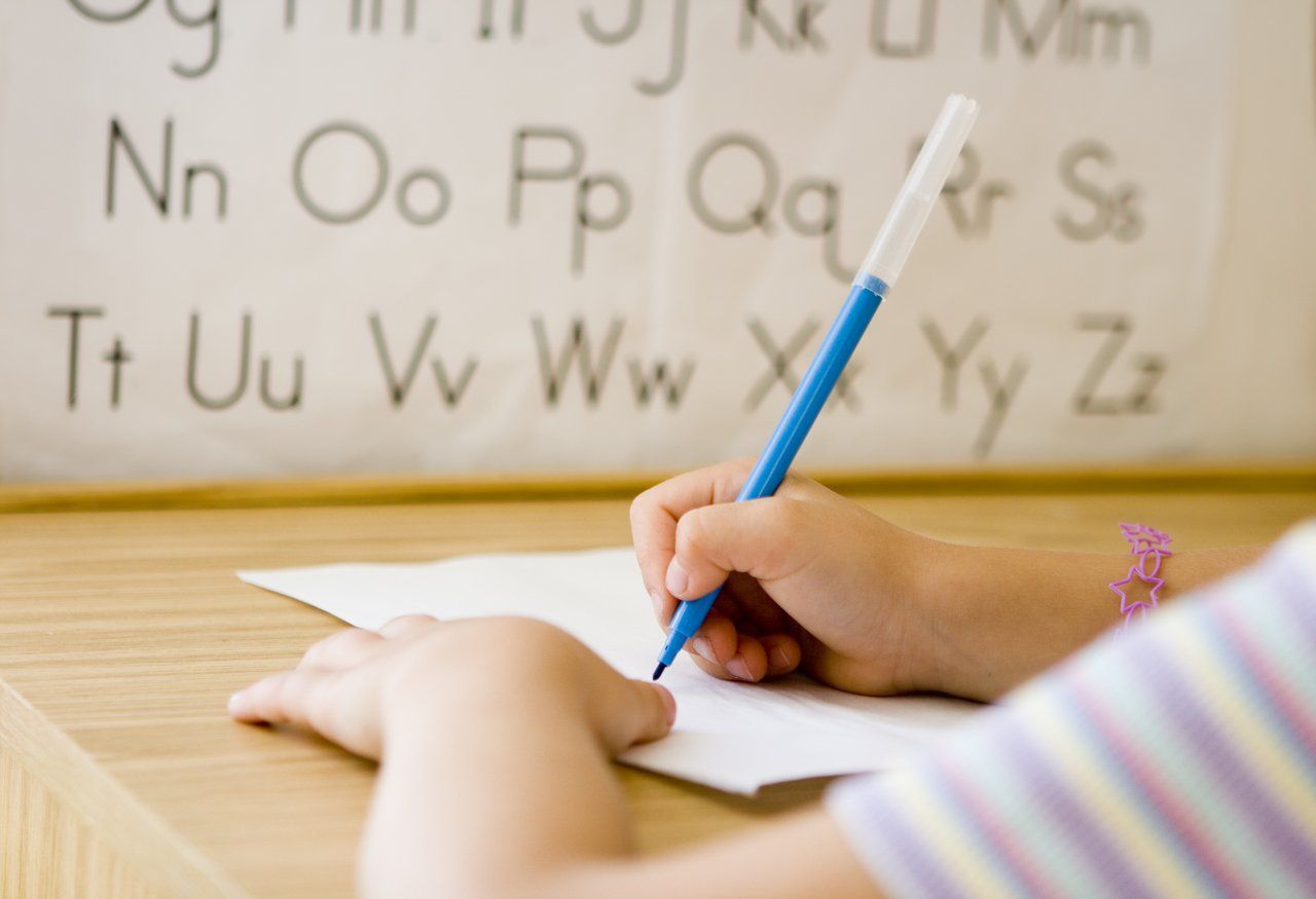 Braço de criança aparece escrevendo em cima de uma mesa com o alfabeto ao fundo