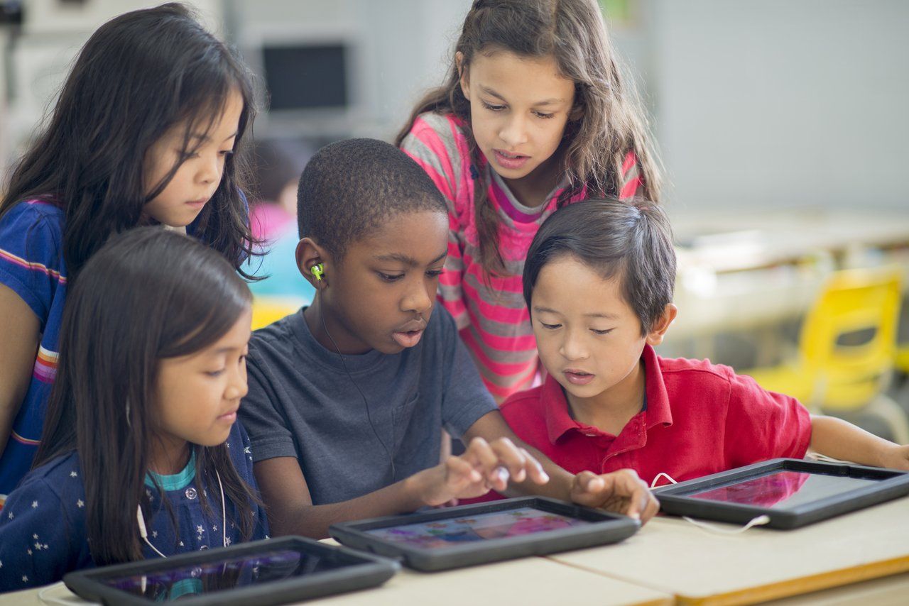 Grupo de alunos reunidos em torno de uma mesa em uma sala de aula brinca com tablets