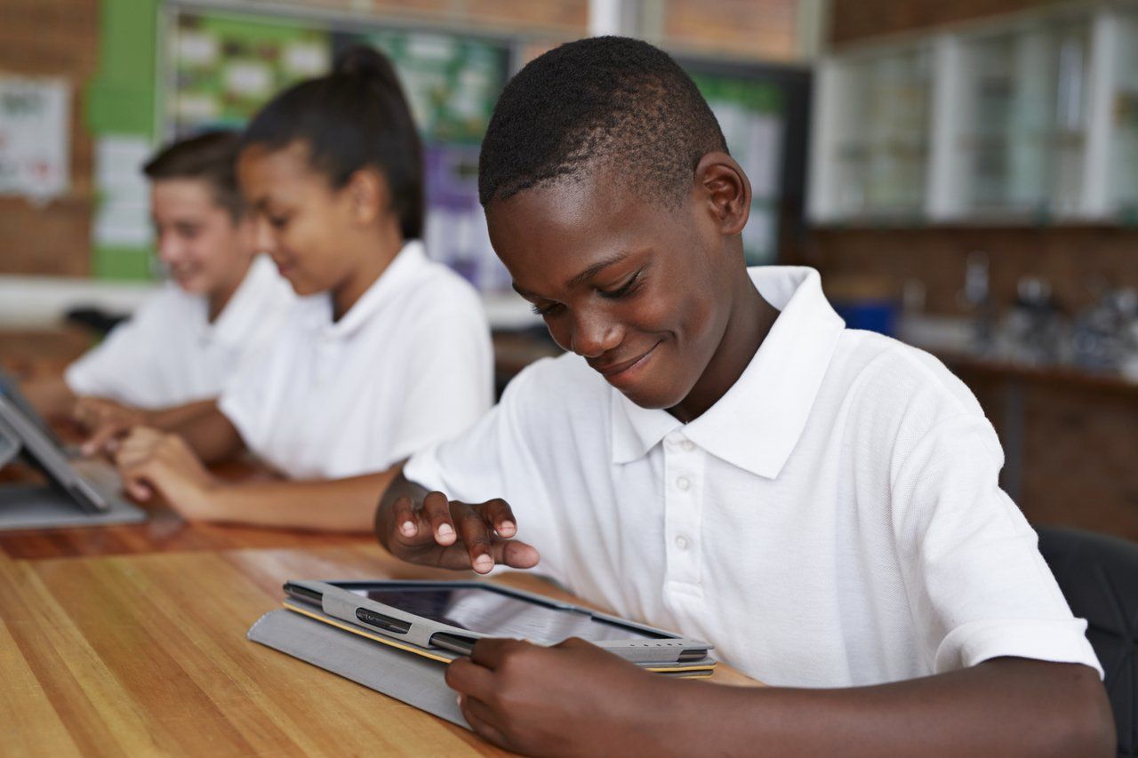 Menino negro sentado em uma mesa escolar com outros estudantes, todos uniformizados com camisas brancas, usa um tablet
