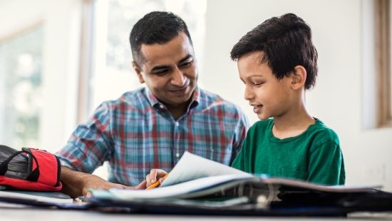 Menino de camisa verde sorri diante de caderno de lição de casa sobre mesa branca, ao lado do pai, que usa camisa xadrez azul e vermelha e também sorri