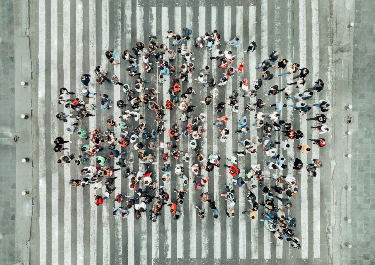 Grupo de pessoas paradas em pé, em cima de uma faixa de pedestres, usando roupas coloridas e vistas de cima, formando a imagem de um balão de fala de histórias em quadrinhos