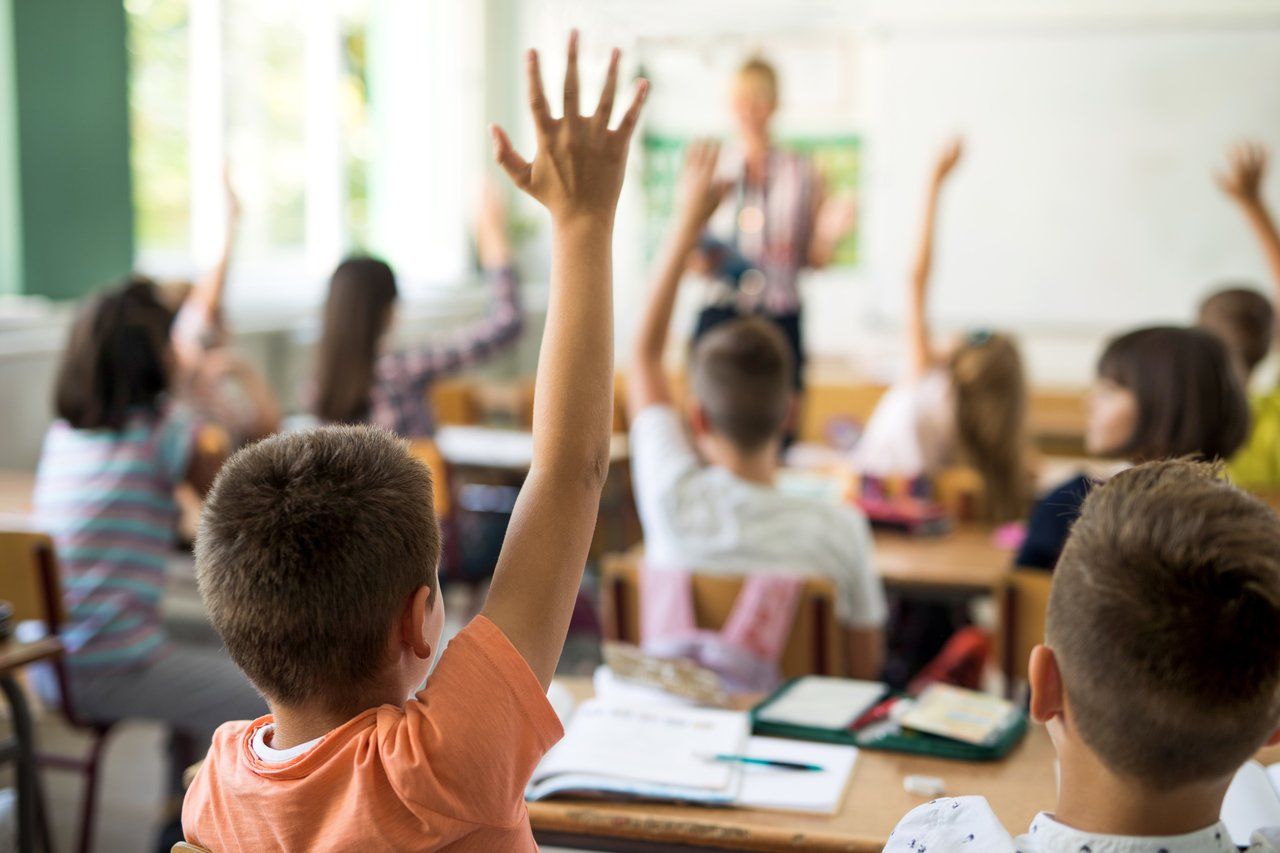 Estudantes levantam as mãos em uma sala de aula, apenas o primeiro está em foco