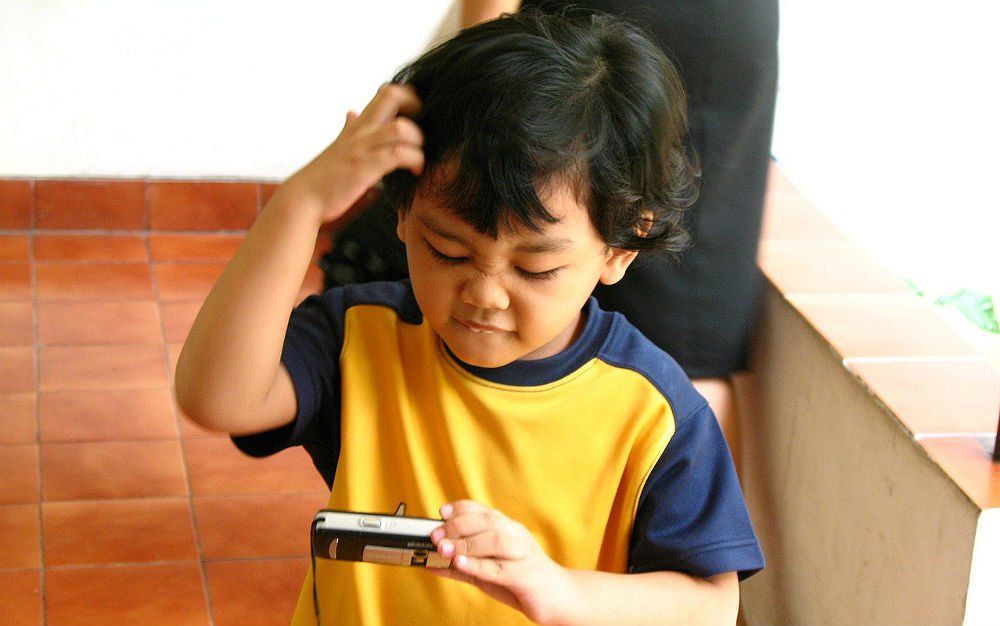 Criança de camiseta amarela e azul olhando para o celular com cara de confusa, coçando a cabeça