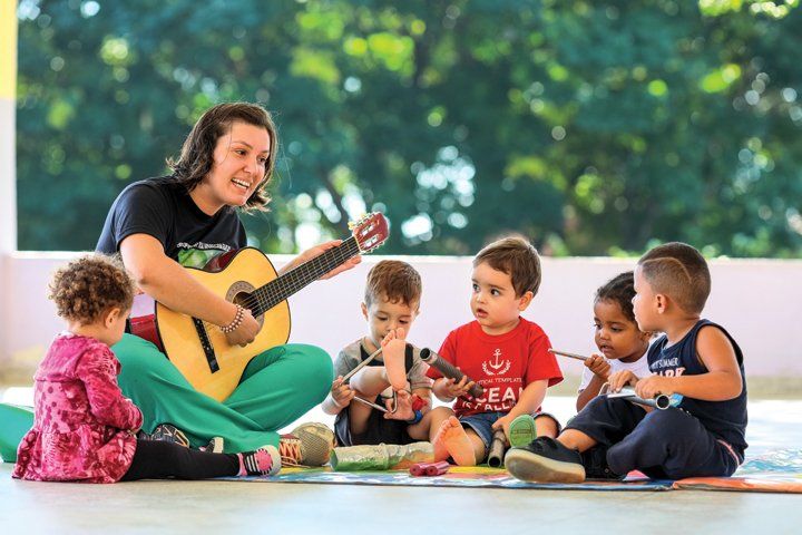 Professora Ariana está sentada em roda com quatro alunos. Ela segura um violão e canta cantigas para os pequenos.