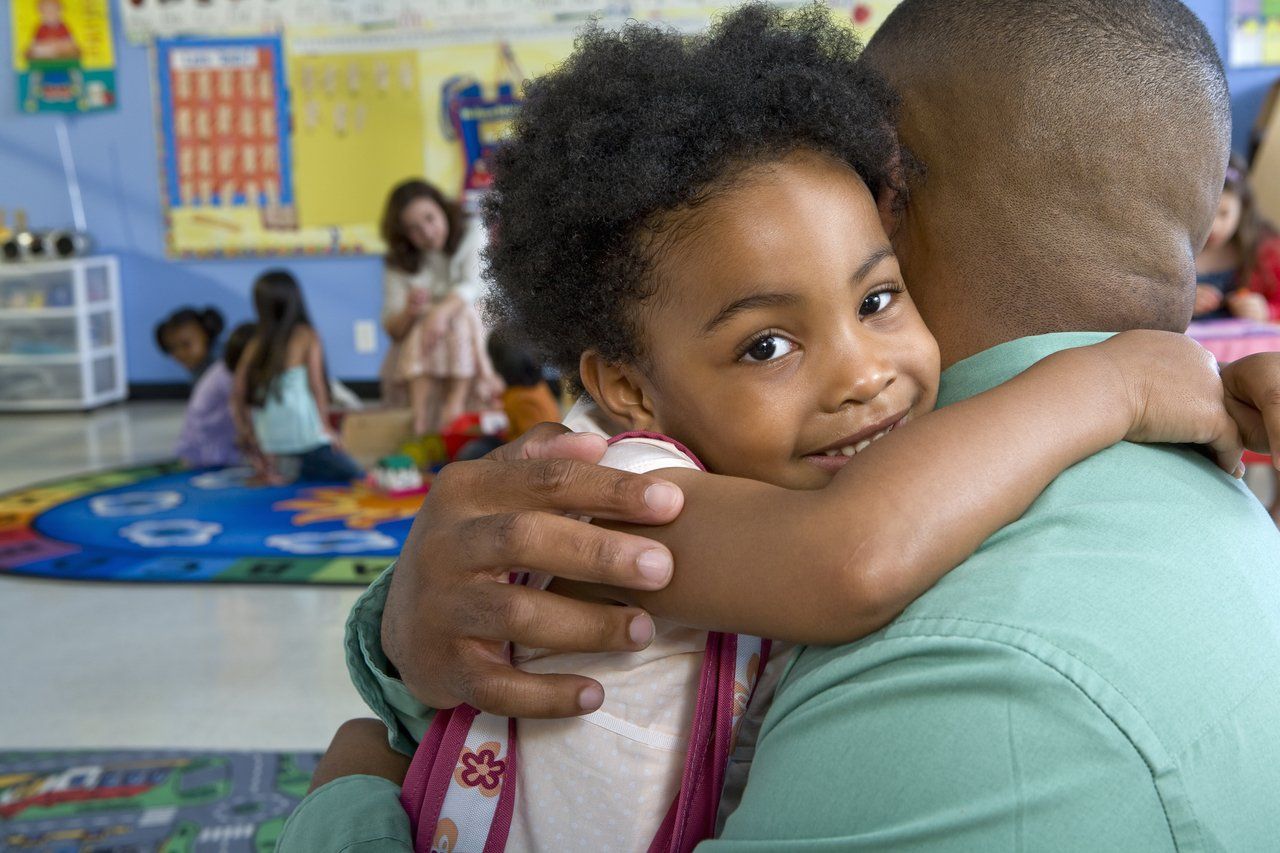 Aluna em primeiro plano olha para a câmera enquanto abraça o pai em uma sala de aula da Educação Infantil