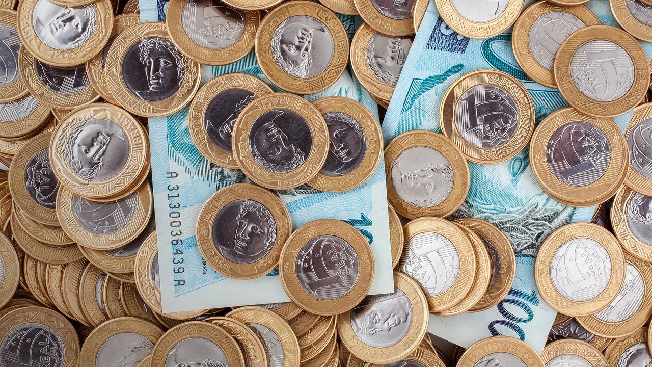 Foto mostra duas notas de cem reais e diversas moedas de um real jogadas sobre uma mesa
