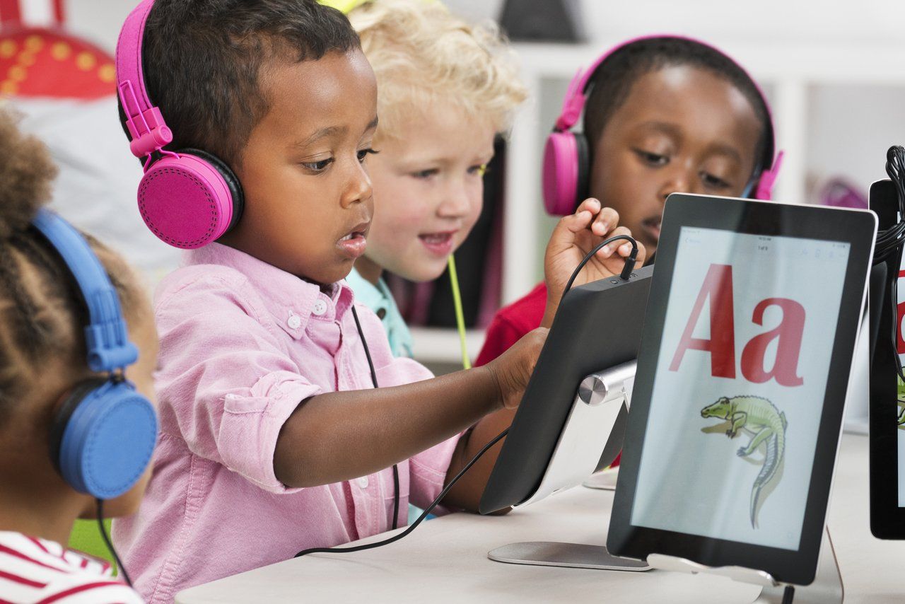 Crianças sentadas em mesas de escola e usando fones de ouvido brincam com tablets que exibem letras coloridas