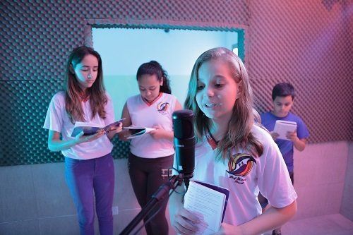  Num estúdio de rádio, uma menina de cerca de 13 anos está a frente do microfone segurando um livro fino. Outras duas estudantes e um estudante estão atrás lendo o mesmo livro.