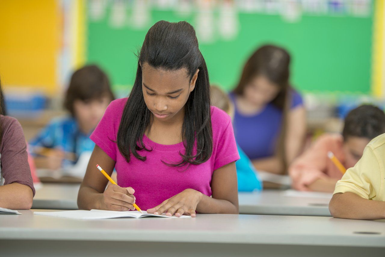 Aluna vestindo blusa rosa escreve em um papel com um lápis laranja, sentada em uma sala de aula