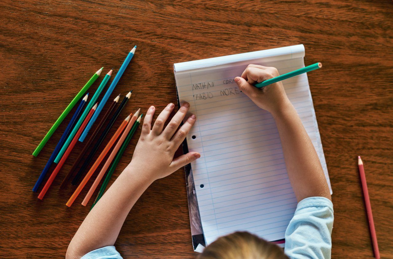 Imagem de cima de criança apoiada em mesa de madeira com caderno aberto e dois nomes escritos com lápis coloridos espalhados ao redor do caderno