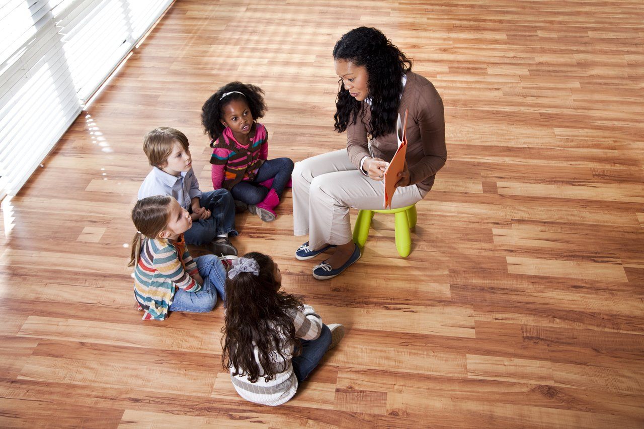 Crianças sentadas em uma roda em um chão de madeira de uma sala espaçosa prestam atenção à professora