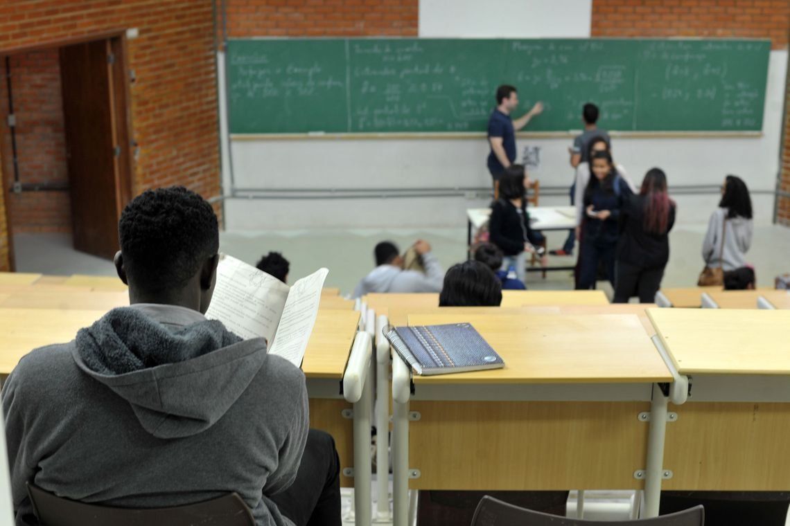 Jovens em uma sala de aula com várias cadeiras vazias e uma lousa com várias inscrições, um jovem negro é visto de costas em primeiro plano