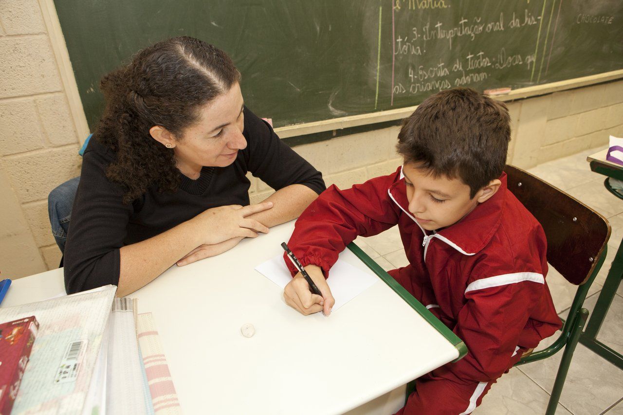 Professora está sentada à mesa com um aluno. Ela olha para ele, enquanto ele escreve uma lista de palavras durante uma sondagem (Crédito: Mariana Pekin)