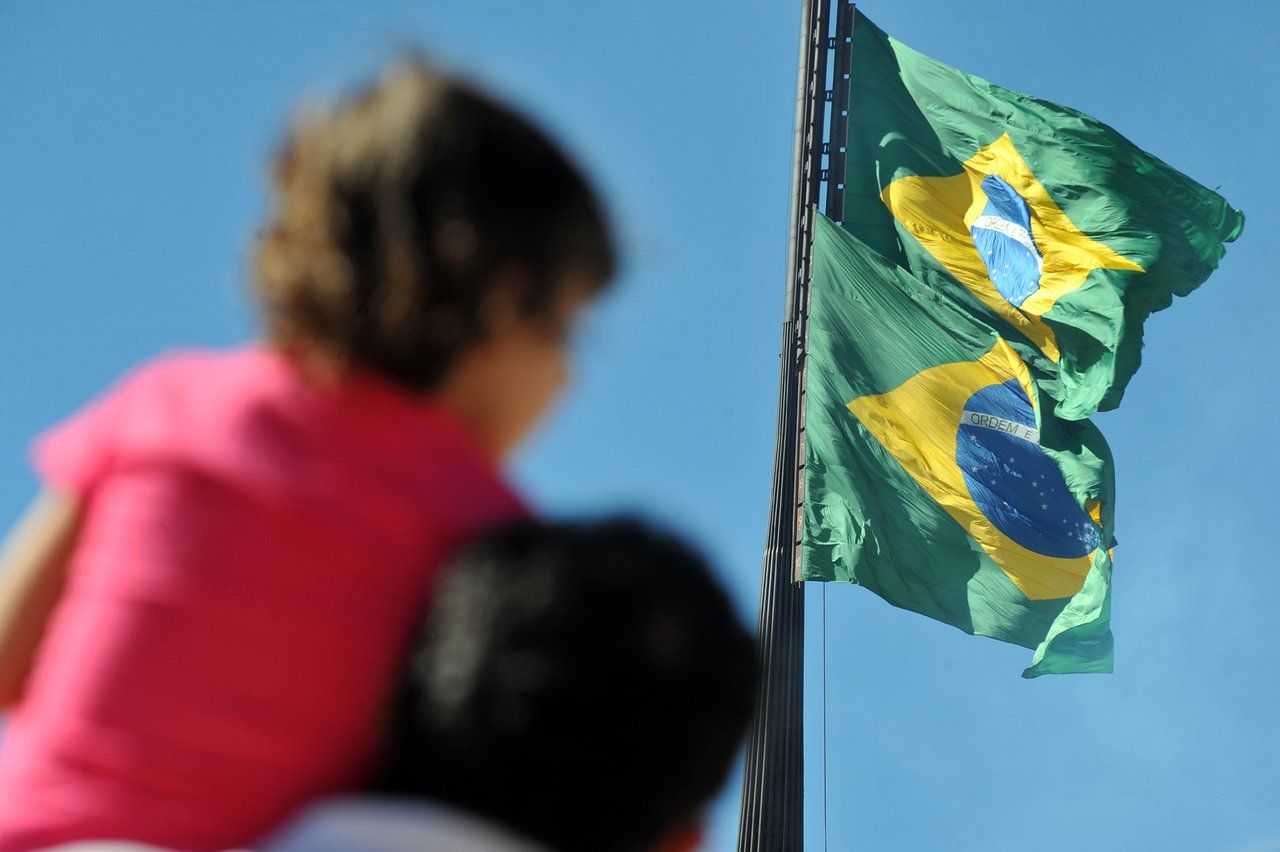 Pai segura menino nos ombros durante comemoração cívica com duas bandeiras do Brasil gigantes hasteadas ao fundo