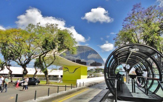 Museu Oscar Niemeyer, em Curitiba (capital do Paraná) aos fundos sob céu azul. Na frente dele, uma árvore com folhas verdes e um ponto de ônibus em forma de tubo, famoso pela cidade.
