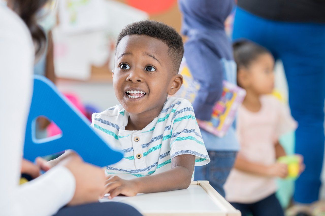 Professora vista apenas de costas entrega uma letra A em azul para criança em sala de aula