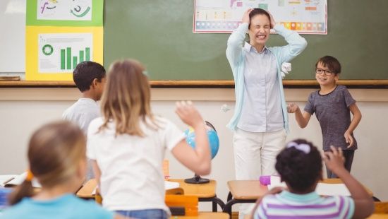 Cinco crianças bagunçam na sala de aula ao redor da professora, de casaco azul e camisa branca. que está de cabelo preso, olhos fechados e mãos tapando os ouvidos