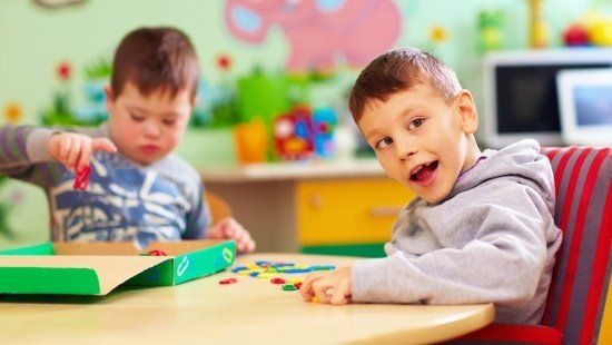 Duas crianças brincam em uma mesa com jogos e brinquedos. Em primeiro plano, uma criança com deficiência intelectual olha em direção ao usuário e sorri. Em segundo plano, um pouco mais desfocada, uma criança com síndrome de Down concentra-se em seu brinquedo.