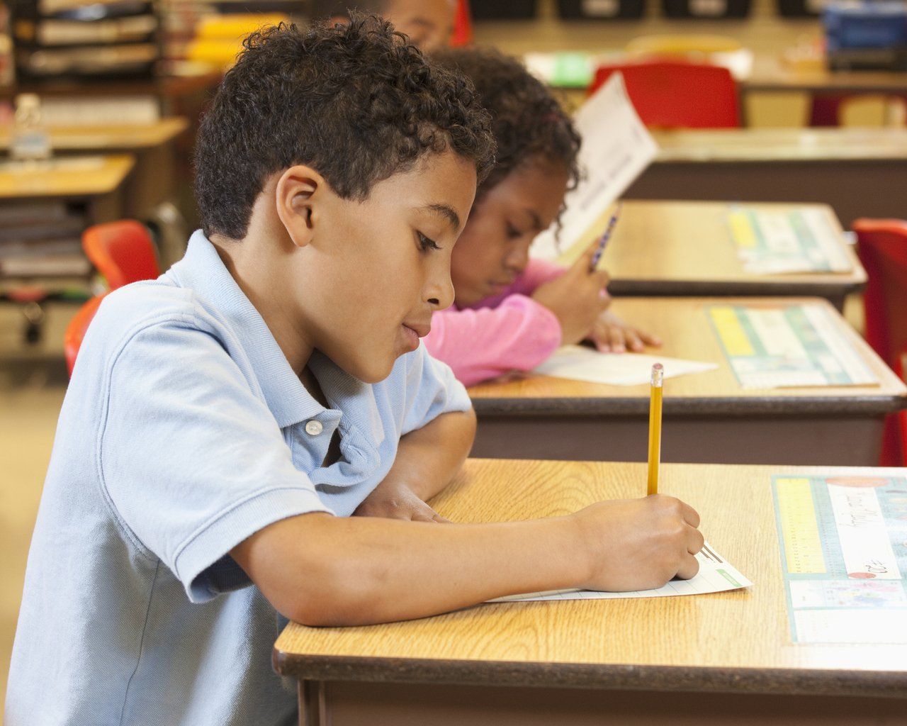 aluno de camisa azul sentado diante de mesa com lápis na mão escrevendo em uma folha de papel 