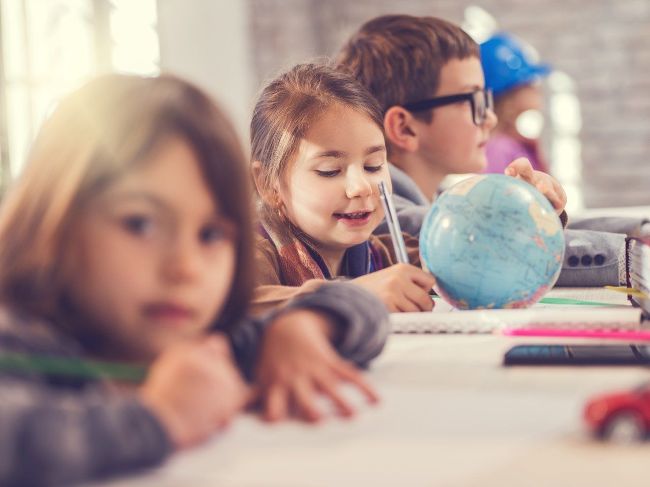 Menina escreve em uma mesa junto com outras crianças tendo um pequeno globo terrestre à sua frente