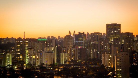 Entardecer na zona sul de São Paulo, de céu laranja sobre uma grande massa de prédios em sua maioria brancos. É possível notar um pedaço da ponte estaiada, um dos símbolos recentes da cidade.