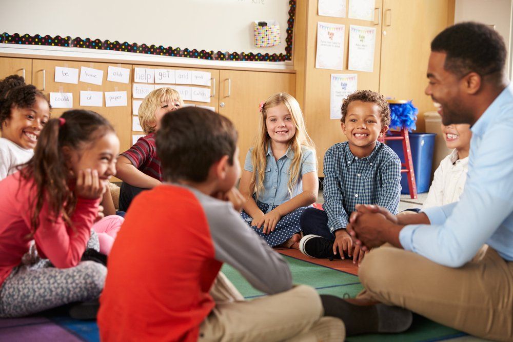 Interior de uma sala de aula. Sete crianças de cerca de 7 anos e um professor negro com cerca de 35 anos estão sentadas em círculo no chão. Todos estão rindo. 