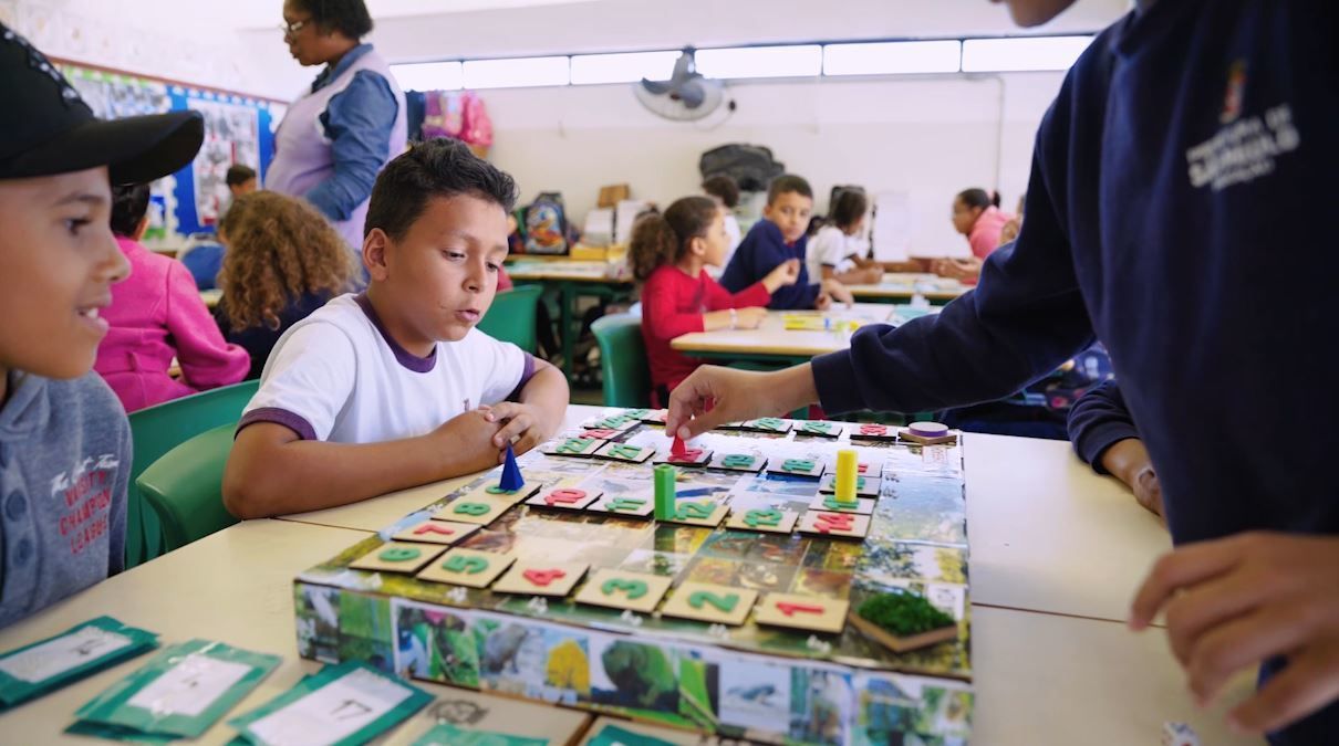 Alunos do 3º ano da EMEF Desembargador Teodomiro Toledo Piza, em São Paulo, brincam com o jogo de tabuleiro Trilha dos Seres Vivos em uma sala de aula divididos em grupos de quatro